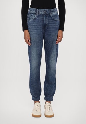 Pantalon jogger en denim bleu foncé avec des poignets élastiques, porté avec des baskets blanches et un haut noir à manches longues, vue de face.