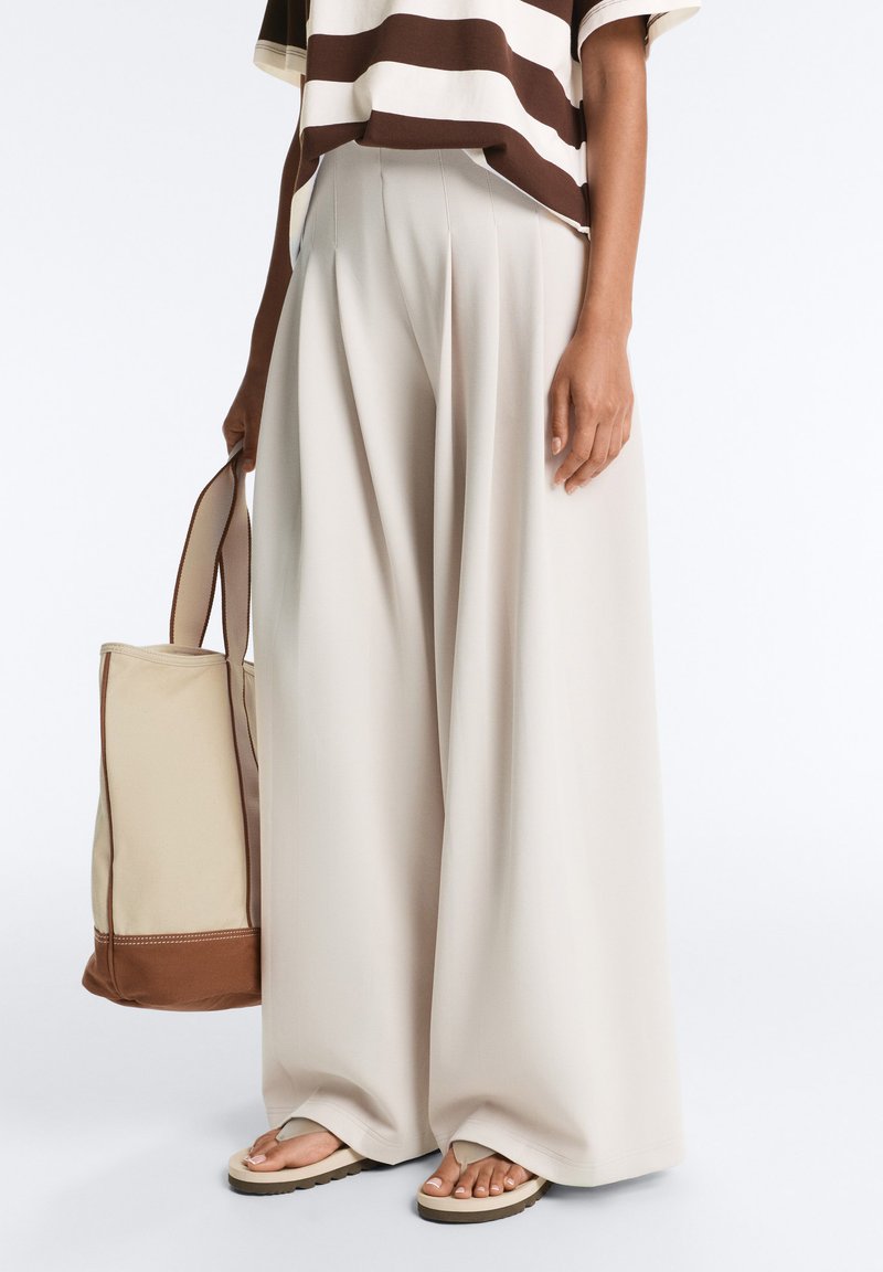 Personne portant un pantalon beige à jambes larges, un haut rayé marron et blanc, tenant un sac fourre-tout beige avec des touches marron, et portant des sandales beiges.
