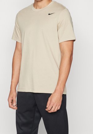 Mężczyzna w beżowej koszulce Nike z krótkim rękawem i czarnym logo Swoosh przy kołnierzu oraz w czarnych spodniach, stojący na jednolitym tle.