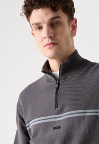 Sivasta zip-up pulover s teksturirano tkanino, s črnim zadrgo in modrim prugastim detajlom čez prsi. Logo "BOSS" na sredini.