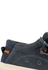 Baskets bleu marine foncé avec une tige en mesh texturé, logo orange et talon renforcé. La semelle est de couleur blanc cassé avec des détails de coutures visibles.