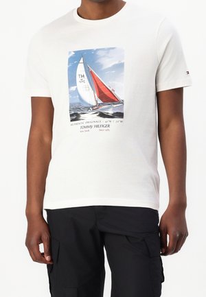 Man draagt een wit T-shirt met een kleurrijke zeilbootafbeelding en tekst, gecombineerd met zwarte cargobroek.
