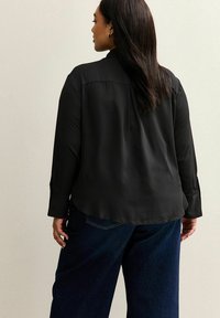 Woman with long dark hair wearing a black long-sleeve blouse and dark blue jeans, standing with her back to the camera.