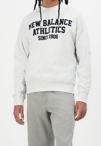 Ljust grå sweatshirt med marinblå text "NEW BALANCE ATHLETICS SINCE 1906." Har lång ärm och ribbade muddar och fåll. Bärs med grå jeans.