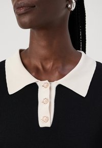 Top nero in maglia con colletto crema e quattro bottoni decorativi, con centro in perla e dettagli dorati. Texture liscia con design aderente.