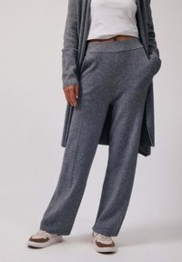 Personne portant un pantalon gris à jambes larges, un cardigan long assorti, un haut court blanc et des baskets blanches avec des détails marron, debout les mains dans les poches.