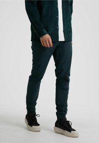 Mörkgrön kordjacka med knappar över en vit T-shirt, kombinerad med mörkgröna byxor och svarta sneakers med krämfärgade sulor.