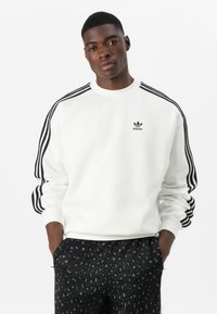 Bela športna majica s črnimi treh črtami na rokavih, z malim črnim logotipom Adidas. Usklajena z črnimi hlačami z vzorcem.