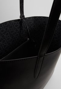 Černá kožená taška přes rameno s texturovaným povrchem, hladkými držadly a vzorovanou černou podšívkou. Obsahuje zipovou kapsu.