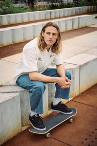 Νεαρό άτομο με μακριά μαλλιά κάθεται σε τσιμεντένιο παγκάκι έξω, ακουμπώντας τα πόδια σε ένα σκέιτμπορντ, φοράει λευκό πουκάμισο και μπλε τζιν.