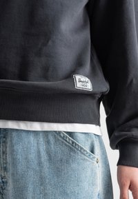 Sudadera gris oscuro con puños y dobladillo acanalados, con una etiqueta de marca blanca. Combinada con unos jeans de denim azul claro, que muestran detalle de los bolsillos.