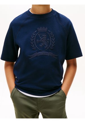 Enfant portant une chemise bleu marine à manches courtes avec un blason comprenant un lion, des branches de laurier et une couronne, assortie à un pantalon vert olive.