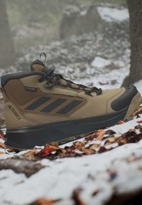 Brauner und schwarzer Wanderschuh mit Schnürsenkeln auf verschneitem Waldboden mit gefallenen Blättern.