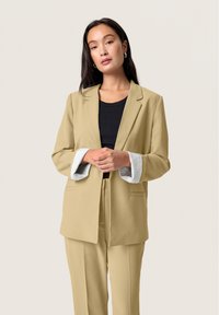 Beige blazer with a tailored fit, featuring striped inner cuffs, paired with a black ribbed top. Smooth fabric, minimalistic design.