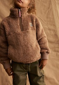 Pullover in pile marrone con colletto alto, chiusura a mezza zip e logo ricamato. Presenta polsini a costine e una vestibilità rilassata. Indossato con pantaloni cargo verdi.