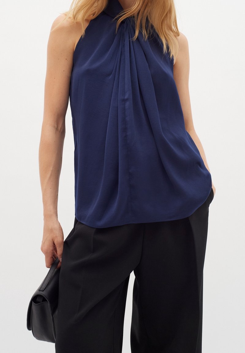 Mörkblå ärmlös blus med plisserad framsida, mjukt tyg, hög halsringning och avslappnad passform, kombinerad med svarta vida byxor.