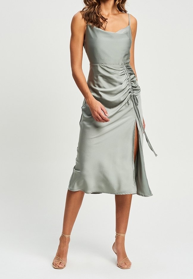 Femme portant une robe midi en satin gris clair avec bretelles fines, détail froncé sur le côté, fente haute à la cuisse et talons beiges à nouer à la cheville.