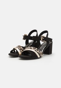 Sandales à talons en noir et imprimé léopard avec un talon bloc, présentant un accent doré brillant et une sangle cheville ajustable. Matériau en daim doux.