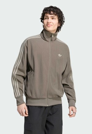 Joven con cabello rizado que lleva una chaqueta de chándal gris de Adidas con cremallera y tres rayas blancas en las mangas, y pantalones negros.