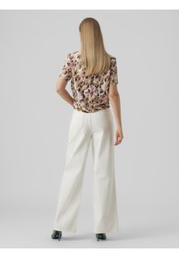 Donna con lunghi capelli biondi che indossa una blusa floreale a maniche corte, pantaloni bianchi a gamba larga e scarpe col tacco teal, in piedi di spalle.