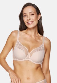 Biustonosz w miękkim beżowym materiale z przezroczystymi koronkowymi detalami, wsparciem z fiszbinami i regulowanymi ramiączkami. Zapięcie z przodu z dekoracyjnymi akcentami.