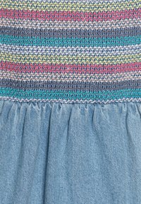 Raidallinen kangastoppi, jossa on monivärisiä vaakaraitoja punaisena, sinisenä, vihreänä ja keltaisena; farkkukuosilla varustettu röyhelöhame vaaleansinisessä sävyssä.