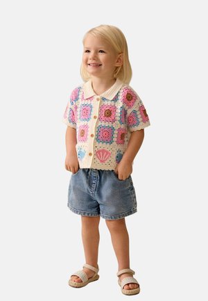 Małe dziecko o blond włosach, ubrane w kolorową szydełkową koszulę na guziki, dżinsowe szorty i sandały, stoi z rękami w kieszeniach, uśmiecha się.