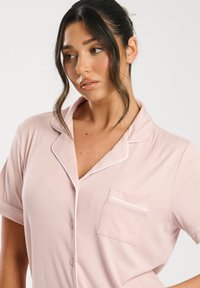 Maglietta da pigiama rosa chiaro a maniche corte in tessuto a coste, con piping bianco lungo il colletto e la tasca. Due bottoni anteriori.