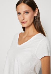Weißes V-Ausschnitt-T-Shirt aus einem weichen, leichten Stoff. Mit kurzen, umgeschlagenen Ärmeln und einem lockeren Schnitt, versehen mit subtiler Textur.