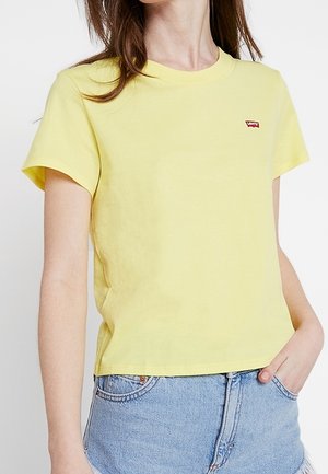 Woman wearing a plain yellow short-sleeve t-shirt and light blue denim shorts with hand in pocket, facing slightly left.