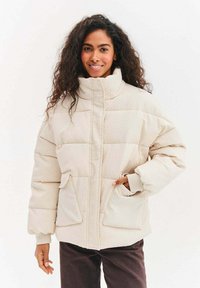 Světlá béžová péřová bunda s prošívanou strukturou, vysokým límcem, dvěma předními kapsami a žebrovanými manžetami. Kostkovaný design s volným střihem.