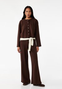 Brun loungewear-set bestående av en croppad knäpp-topp och vida byxor, accentuerad med ett krämfärgat bälte, tillverkat av mjukt, texturerat tyg.