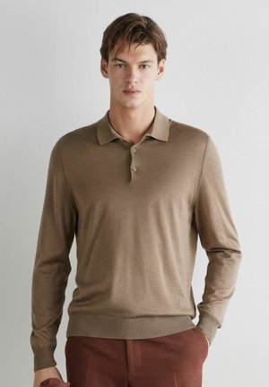 Junger Mann trägt einen hellbraunen Polo-Pullover mit langen Ärmeln und rostfarbene Hose, steht vor einer schlichten weißen Wand.