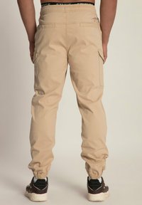 Man met beige cargobroek met elastische boorden en bruine sneakers, weergegeven van achteren tegen een neutrale achtergrond.