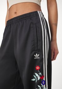 Svarta satin joggers med elastisk midja, med vita sidremsor, blommig broderi och en Adidas-logotyp.
