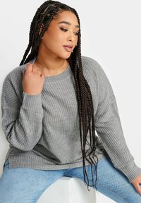 Grå stickad tröja med lös passform, ribbad textur och sidsprund, ihop med ljusblå denimjeans. Lång flätad frisyr.