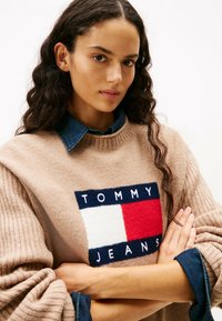 Frau mit lockigem, dunklem Haar, die einen beigen Tommy Jeans Pullover über einem Jeanshemd trägt, mit verschränkten Armen, die in die Kamera schaut, vor einem weißen Hintergrund.