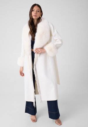Femme portant un long manteau blanc avec col et poignets en fourrure, un jean large sombre et des sandales ouvertes sur un fond gris uni.