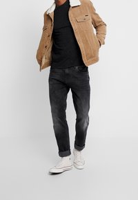 Chaqueta de pana marrón con cuello de sherpa blanco, camiseta negra moteada, jeans desgastados negros y zapatillas altas blancas. Atuendo casual.