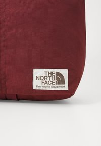 Burgundska tkanina torba s teksturiranim završetkom, koja ima bijelu oznaku s ispisom loga "THE NORTH FACE" i tekstom "Fine Alpine Equipment".