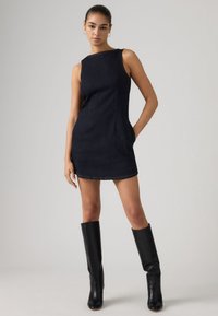 Γυναίκα που φοράει μαύρο μίνι φόρεμα χωρίς μανίκια με λεπτομέρεια σε έναν ώμο και μαύρες δερμάτινες μποτάκια μέχρι το γόνατο, στέκεται με το ένα χέρι στην τσέπη.