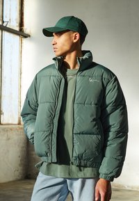 Chaqueta tipo puff verde con cuello alto, dobladillo elástico y bolsillos laterales, usada sobre una camiseta verde a juego, acompañada de una gorra verde.