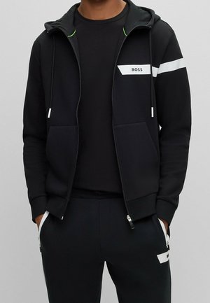 Mand iført en sort hættetrøje med lynlås og matchende joggingbukser med hvid stribe og "BOSS"-logo på brystet og lår-lommerne.