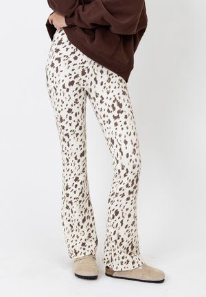 Άτομο που φοράει λευκό παντελόνι με καφέ animal print, μπεζ παπούτσια slip-on και καφέ φούτερ, στέκεται μπροστά σε απλό φόντο.
