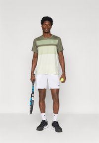 Cămașă sport verde și gri deschis cu dungi orizontale, asociată cu pantaloni scurți albi, încălțată cu adidași negri, ținând o rachetă de tenis și o minge.