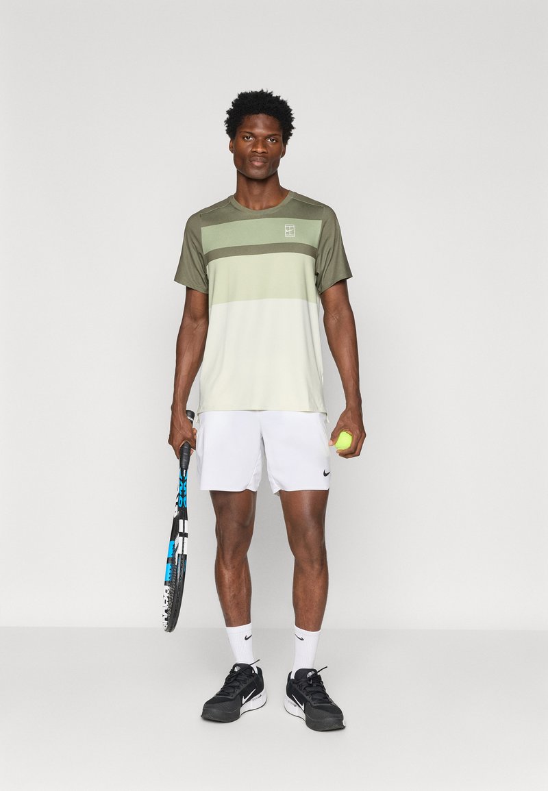 Green and light gray athletic shirt with horizontal stripes, paired with white shorts, black tennis shoes, holding a tennis racket and ball.