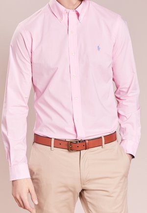 Homme portant une chemise rose clair boutonnée, un pantalon beige et une ceinture en cuir marron avec une boucle dorée, les mains détendues le long du corps.
