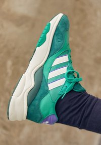 Grön atletisk sneakers med vita ränder, som har en blandning av mocka och syntetiska material, rundad tå och en mörk sula.