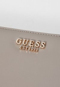 Jasno beżowy portfel o gładkiej fakturze, z wypukłym złotym logo "GUESS" oraz "EST 1981" poniżej. Subtelne detale stębnowania wzdłuż krawędzi.