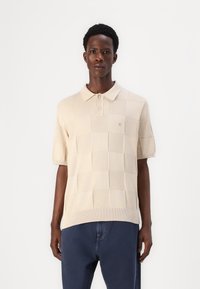 Beige rövidujjú póló, textúrált sakkmintával, két gombos nyakkivágással és logóval a mellkasán. Kék farmernadrággal párosítva.